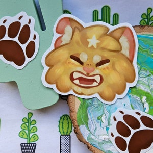 May include: A collection of stickers featuring a cartoon cat with an angry expression, a paw print, and a cactus design. The cat sticker is light brown with a white star on its forehead. The paw prints are brown with white outlines.
