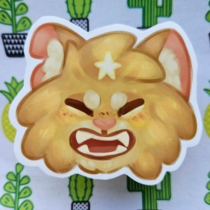 May include: A sticker featuring a cartoon cat with a fierce expression. The cat is primarily yellow and tan, with a white star on its forehead and pink accents in its ears and nose. The sticker has a white border.