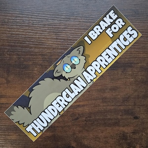 May include: A rectangular sticker with a cartoon cat illustration and the text "I BRAKE FOR THUNDERCLAN APPRENTICES" in white. The cat is light gray with blue eyes and a yellow and black background.