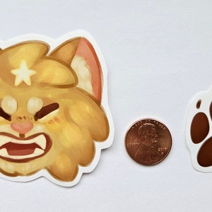 May include: Two stickers: a cartoon cat face with a star and a brown paw print. The cat face is golden yellow with a white star and pink accents. A US penny is next to the stickers for size comparison.