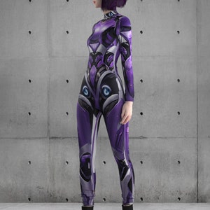 Puede incluir: Una mujer con un mono morado y negro de dise&ntilde;o futurista. El mono tiene un brillo met&aacute;lico y presenta una variedad de formas geom&eacute;tricas y l&iacute;neas. La mujer est&aacute; de pie frente a una pared de hormig&oacute;n gris.