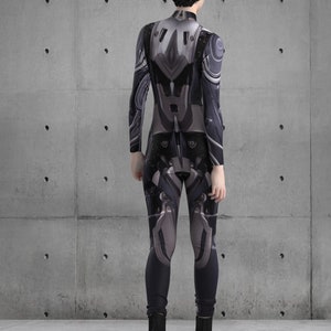 Puede incluir: Un mono de cuerpo entero con un dise&ntilde;o futurista en tonos grises y negros. El traje presenta paneles y detalles detallados, creando una est&eacute;tica tecnol&oacute;gica. El modelo lleva botas negras.