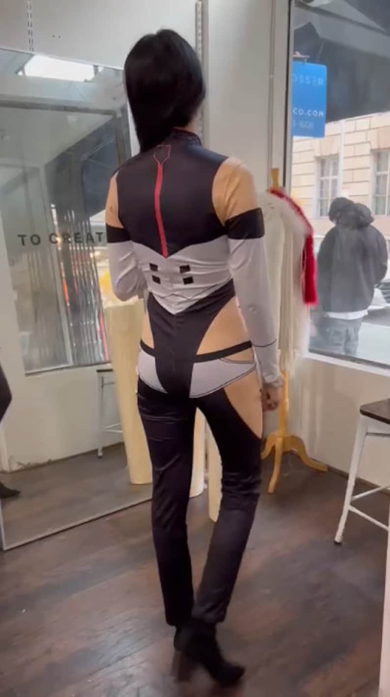 May include: A person wearing a black and white jumpsuit with a red stripe down the back. The jumpsuit has a white panel on the back with a geometric design. The person is standing in a store with a mirror behind them.