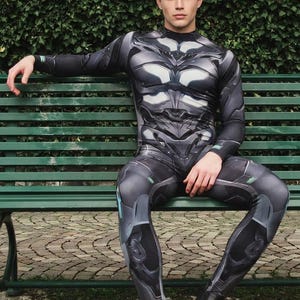 Könnte beinhalten: Ein Ganzkörper-Bodysuit in Grau- und Schwarztönen mit futuristischem Design, langen Ärmeln und weißen und grünen Akzenten. Die Person trägt schwarze Stiefel und sitzt auf einer grünen Bank. Im Hintergrund eine Hecke.
