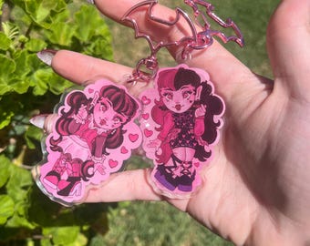May include: Two pink acrylic keychains featuring cartoon characters with dark hair and pink accents. The keychains are attached to a pink bat-shaped keyring. The characters are wearing pink and black outfits.