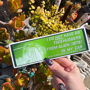 Peut inclure: Un autocollant de pare-chocs vert avec le texte "I DO NOT HAVE 28 FACEHUGGERS FROM ALIEN (1979) IN MY CAR. THE RATTLING IS UNRELATED." L'autocollant présente une image d'un facehugger. L'autocollant est tenu devant un fond de plantes.