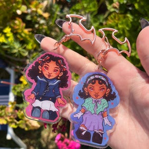 May include: Two acrylic keyrings with cartoon illustrations of girls. One girl wears a navy blue sailor outfit, and the other wears a green and purple outfit. Both keyrings have rose gold bat-shaped clasps.