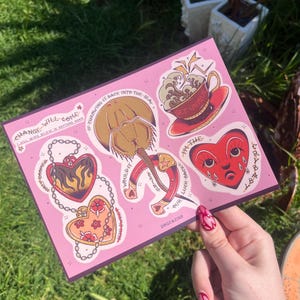 Puede incluir: Una hoja de pegatinas rosa con varios diseños estilo tatuaje. Incluye un corazón con llamas, una taza de té con el texto "GET UNIQUE", una herradura y un corazón llorando. La hoja también tiene texto que dice "CHANGE WILL COME" y "OUR LUCK BACK".