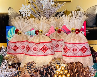 May include: Four burlap gift bags with a red and white snowflake pattern, tied with jute twine and adorned with gold bells and red jingle bells. The bags are surrounded by pine cones and gold ornaments, creating a festive holiday display.