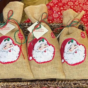 May include: Three burlap gift bags with Santa Claus faces, tied with green and red twine. Each bag has a tag that says "Merry Christmas". The bags are in a woven basket, with a red and white snowflake patterned fabric in the background.