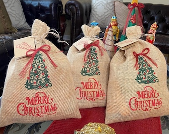 May include: Three burlap gift bags with red bows and tags that read "Merry Christmas." Each bag features a green Christmas tree design and the words "Merry Christmas" in red. The bags are tied with twine.