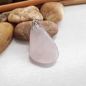 May include: A pale pink rose quartz pendant with a silver bail, resting on a bed of smooth, brown and beige stones. The stones are arranged around a pair of wooden chopsticks.