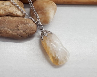 May include: A silver chain necklace with a clear, yellow-tinted gemstone pendant. The pendant is set in a silver frame with intricate details. The necklace is displayed on a white surface with brown stones.