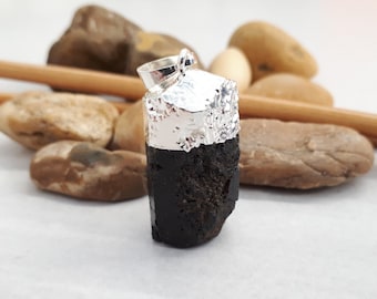 May include: Black gemstone pendant with a silver bail. The pendant is a raw, unpolished stone with a rough, natural texture. It is set against a white background.