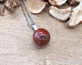 Op de afbeelding: Een zilveren ketting met een ronde, rode edelsteen hanger. De hanger is omringd door gedroogde bloemblaadjes.