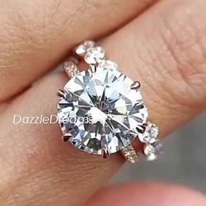 May include: A rose gold engagement ring set with a large, round, clear centre stone and two smaller, round, clear stones on each side. The ring is set on a band with smaller, clear stones.
