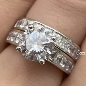 May include: A silver ring set with a large, round, clear cubic zirconia stone surrounded by smaller stones. The ring is worn on a finger with a matching band.
