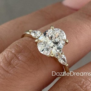May include: A gold engagement ring with a large oval-shaped diamond and smaller pear-shaped diamonds on either side. The ring is set on a finger.
