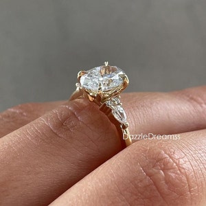 May include: A gold engagement ring with a large oval-shaped center stone and three smaller pear-shaped stones on the band. The ring is set on a finger.