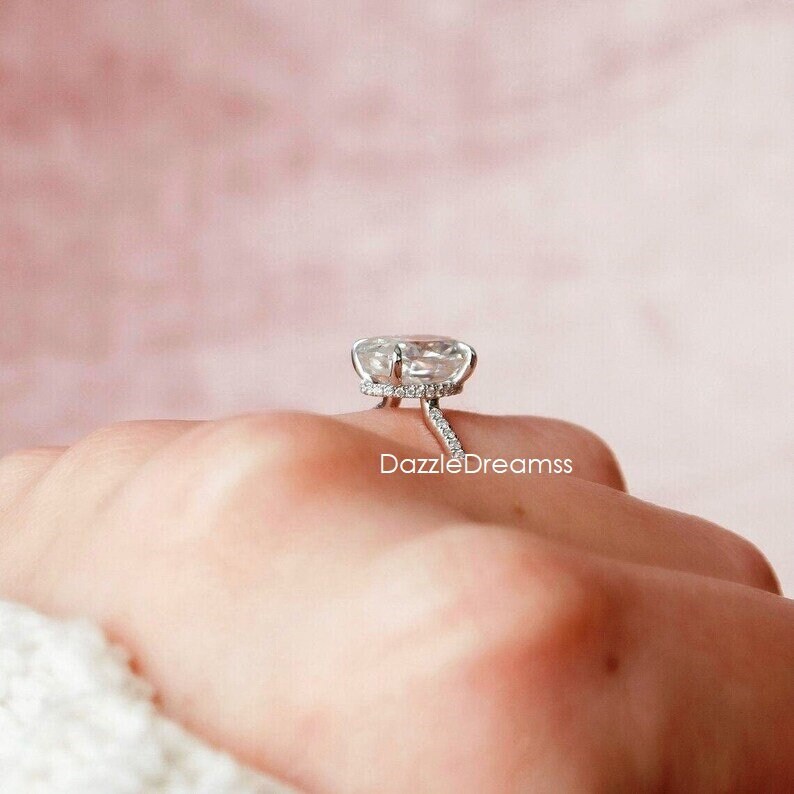 May include: A silver engagement ring with a large, clear, oval-shaped center stone surrounded by a halo of smaller stones. The ring is set on a thin band with a delicate, pave-set band.