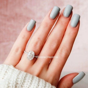 May include: A close-up of a hand wearing a silver ring with a large oval-shaped diamond. The hand is painted with a light gray nail polish.