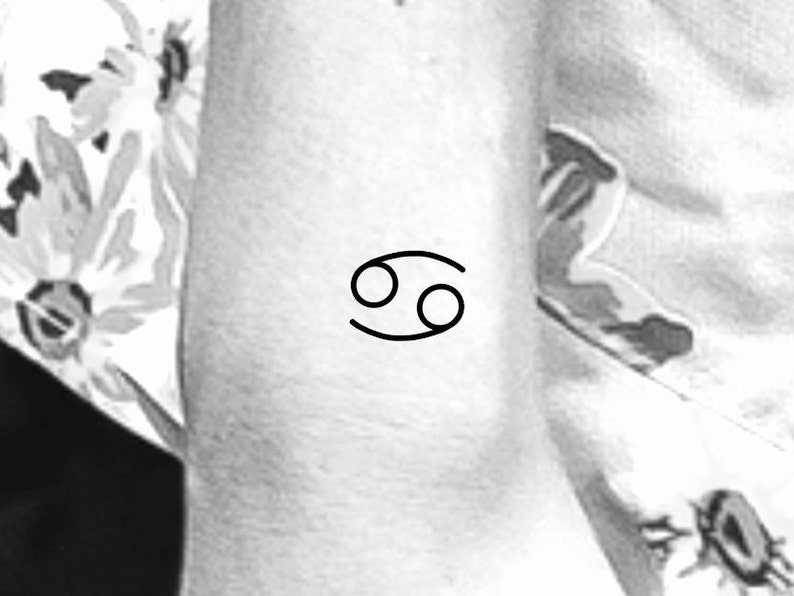 May include: A black line drawing of the Cancer zodiac symbol on a person's arm.