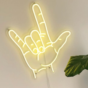 May include: A neon sign in the shape of a hand making the American Sign Language symbol for "I love you". The sign is lit up in a bright yellow color and is mounted on a white wall. A green leafy plant is in the background.