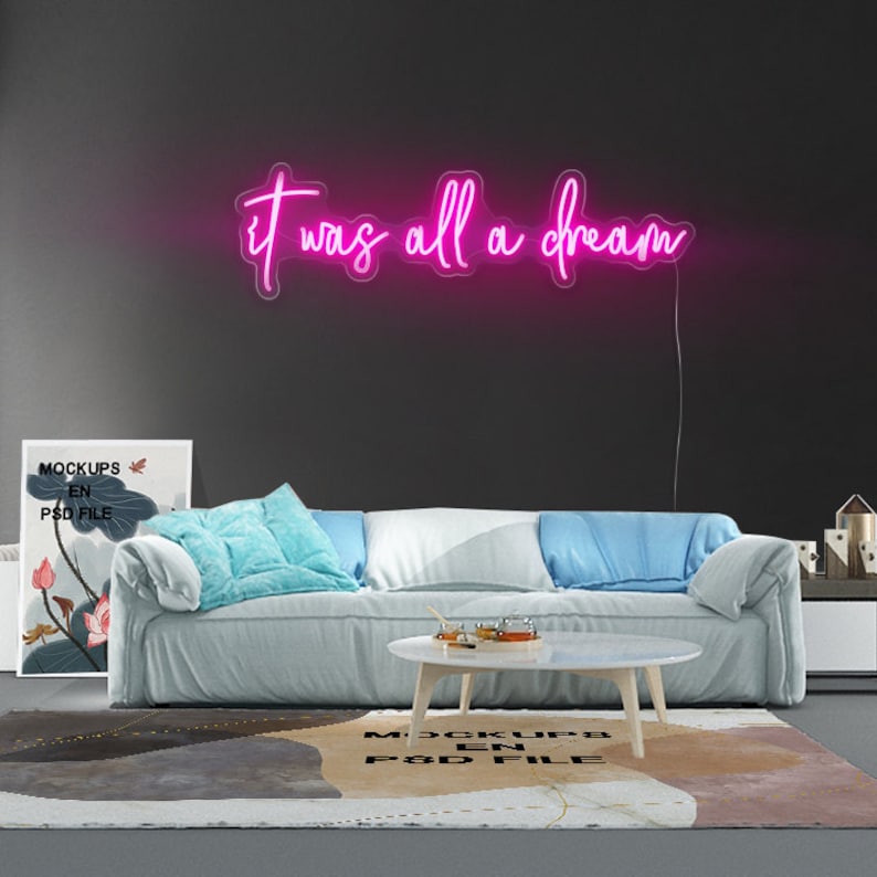 Puede incluir: Un letrero de ne&oacute;n rosa con el texto "it was all a dream" colgado en una pared gris oscuro sobre un sof&aacute; blanco con almohadas azules. El sof&aacute; est&aacute; frente a una alfombra gris con un patr&oacute;n blanco y negro.