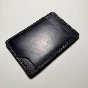 Puede incluir: Un tarjetero de cuero negro con un diseño minimalista. La cartera presenta un acabado liso y mate y costuras visibles alrededor de los bordes. Tiene un bolsillo en un lado y una abertura curva en el otro.