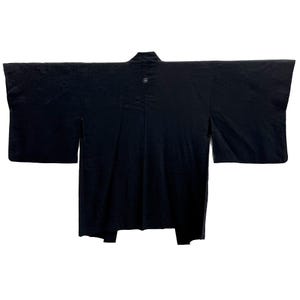 May include: A black kimono with wide sleeves, displayed flat. The garment has a subtle textured pattern and a small circular emblem near the collar. The kimono is made of a dark fabric, likely silk or a similar material.