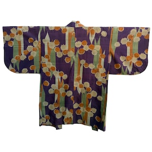 Op de afbeelding: Een vintage kimono in een levendige paarse kleur, versierd met een herhalend bloemmotief in oranje, crème en groen. Verticale strepen voegen een contrast toe. De kimono wordt plat gepresenteerd, waarbij de traditionele vorm en stijl worden getoond.