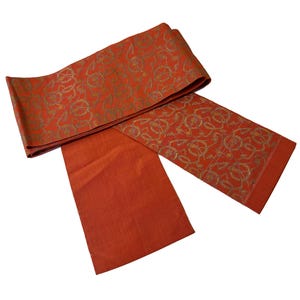 May include: A burnt orange obi belt with gold-colored decorative patterns. The belt is folded over itself, with one end extending downward. The decorative pattern is repeated on the top and bottom of the belt.