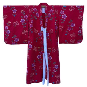 May include: A red kimono with a floral and butterfly pattern. The kimono features purple and white flowers, green stems, and white butterflies. A white sash is tied at the waist. The kimono is approximately 55 inches long.
