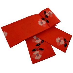 Puede incluir: Tela naranja brillante con un estampado floral. Las flores son blancas, rosas y grises con tallos y hojas negras. La tela está doblada, posiblemente un obi o cinturón decorativo.