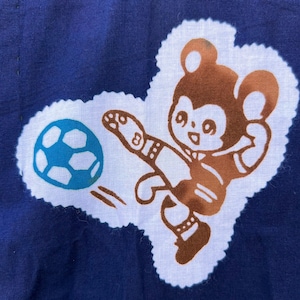 May include: A navy blue fabric with a white, cloud-shaped patch featuring a brown cartoon bear kicking a blue and white soccer ball. The bear is wearing a soccer uniform.