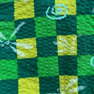 May include: Green and yellow checkered fabric with a white floral pattern. The fabric has a textured surface.