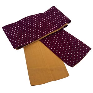 Puede incluir: Un cinturón obi burdeos y dorado con un patrón de cuadrados blancos. El obi está doblado sobre un trozo de tela rectangular dorado. El obi es un cinturón japonés tradicional que se usa con un kimono.