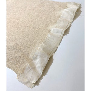 May include: A close-up of a cream-colored woven fabric with a frayed edge. The fabric has a textured surface and a subtle sheen.
