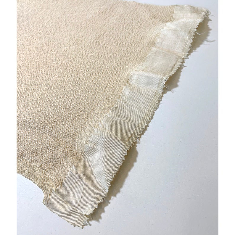May include: A close-up of a cream-colored woven fabric with a frayed edge. The fabric has a textured surface and a subtle sheen.