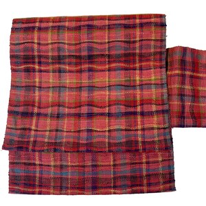 May include: Red, blue, and yellow plaid fabric with a woven texture. The fabric is folded in half and the edges are frayed.