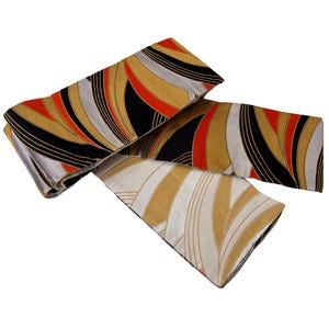 May include: Three decorative fabric pieces with a repeating wave pattern in black, gold, red, and silver. The fabric is folded and layered, creating a dynamic visual effect. The design is reminiscent of Art Deco style.
