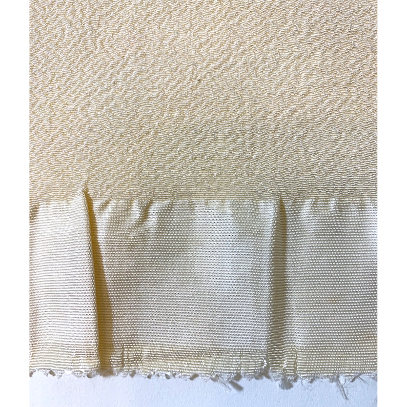May include: A close-up of a cream-colored fabric with a textured pattern. The fabric has a smooth, silky feel and a subtle sheen. The fabric is folded in half, revealing a slightly different texture on the underside.