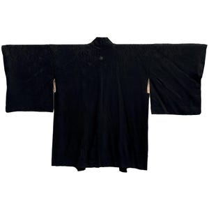 Puede incluir: Un kimono negro con un sutil patrón geométrico. La prenda tradicional japonesa presenta mangas anchas y cuello mandarín. La tela tiene un ligero brillo y se puede ver un pequeño botón decorativo cerca del cuello.