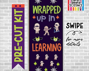 Op de afbeelding: Een paarse rechthoekige deurdecoratieset voor in de klas met de tekst WRAPPED up in LEARNING in limoengroen en oranje. Het ontwerp bevat cartoonmummies, limoengroene vleermuizen, witte spinnenwebben en gele sterren op een donkerpaarse achtergrond.