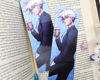May include: Two anime-style bookmarks showing a character with white hair and a dark outfit, holding a coffee cup. The bookmarks are placed on an open book.