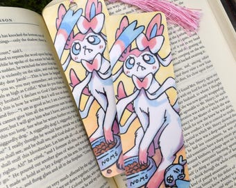 May include: Two illustrated bookmarks featuring a Sylveon-like character with pink, blue, and white accents, holding a bowl of food labelled "NOMS". The bookmarks have pink tassels and are set on an open book.