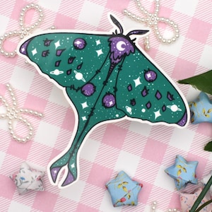 Puede incluir: Una pegatina caprichosa con una polilla verde con un diseño celestial. Las alas de la polilla están adornadas con planetas, estrellas y detalles morados. La pegatina está sobre un fondo a cuadros rosa y blanco, con elementos decorativos.