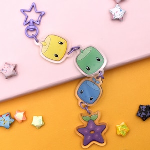 May include: A purple star-shaped keychain with a lobster clasp and a chain of colorful, cartoon-style charms. The charms include a yellow square, a green square, a blue square, and a purple star. Decorative origami stars are scattered around the keychain.