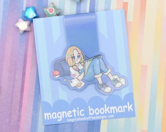 May include: A magnetic bookmark with an anime-style illustration of a person with blonde hair, wearing blue overalls, and trainers, lying down and drawing. The bookmark has the text "magnetic bookmark" and a website address.