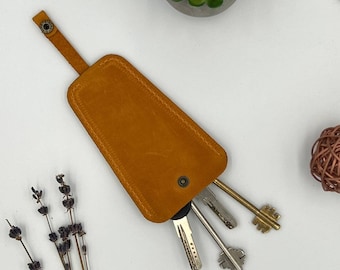 May include: A mustard-colored leather key holder with a snap closure and a loop. Several keys of different shapes and sizes are partially visible. The key holder is on a white surface, with a small succulent plant and decorative items in the background.